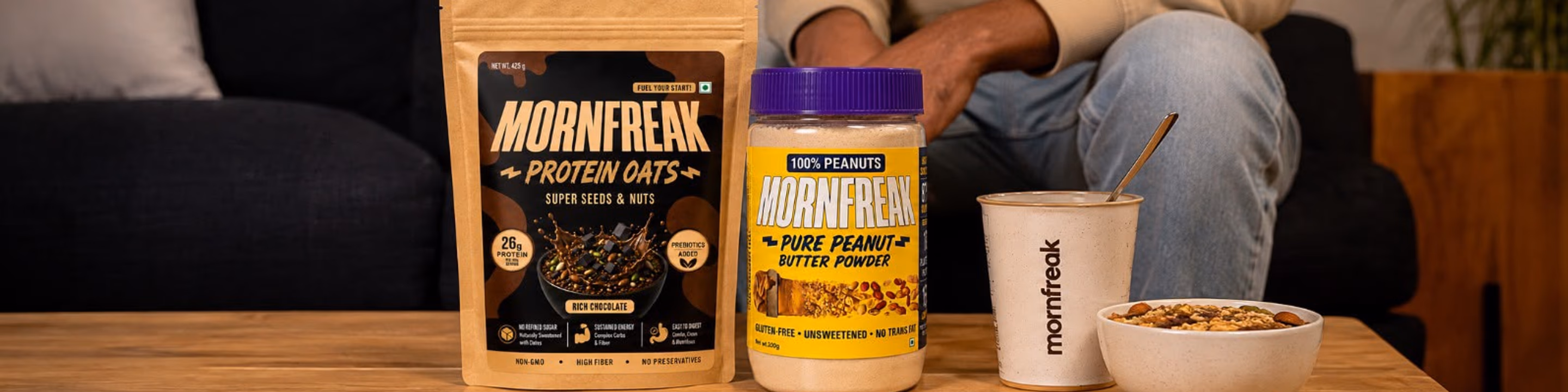 Mornfreak Protein Oats pouch, peanut butter powder, branded cup, and a prepared bowl on a wooden table